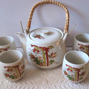 May include: A white ceramic teapot with a woven handle and four matching teacups. The teapot and cups feature a floral and bamboo design in shades of red, gold, and green.