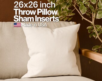May include: A white 26x26 inch throw pillow sham insert made in the USA. The pillow is sitting on a beige couch with a wooden armrest.