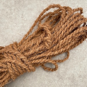 Peut inclure: Un paquet de corde épaisse en fibre naturelle marron. La corde est torsadée et semble être faite d'un matériau grossier. La corde est regroupée, avec des boucles visibles en haut. Le fond est une surface texturée gris clair.