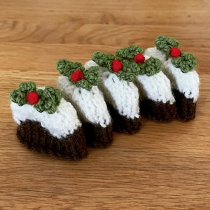 May include: Five knitted Christmas pudding ornaments with green holly and red berries on top. The ornaments are arranged in a row on a wooden surface.