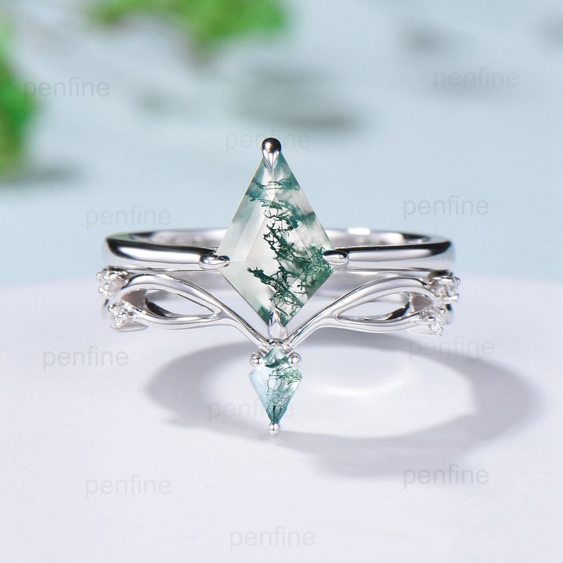 May include: A silver ring with a green moss agate gemstone in a kite shape. The ring has a delicate, intricate design with two bands and a smaller green moss agate gemstone hanging below the main stone.