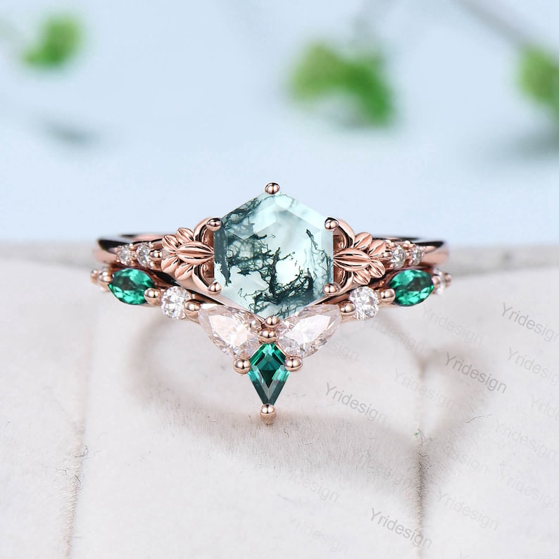 May include: A rose gold engagement ring set with a hexagonal green moss agate center stone. The ring is accented with smaller green emerald stones and white diamonds. The band is adorned with delicate floral details.