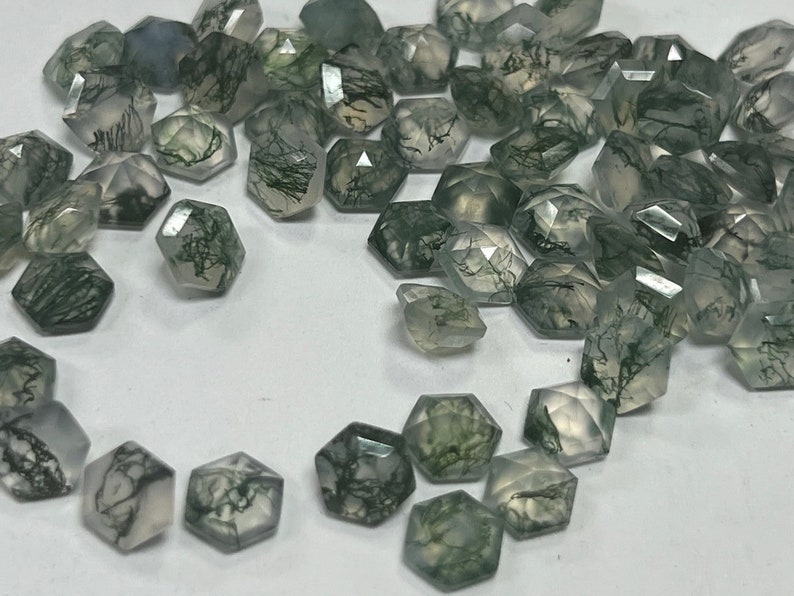 May include: A collection of green and white faceted gemstone beads. The beads are in the shape of a hexagon and have a natural, cloudy appearance. The beads are scattered on a white surface.
