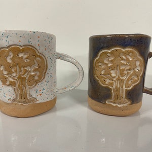 May include: Two ceramic mugs with a tree design. One mug is white with blue and orange speckles, the other is dark blue. Both mugs have a tan tree design and a tan base. The mugs have a curved handle.