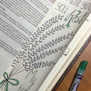Può includere: Una pagina della Bibbia con un'illustrazione disegnata a mano di un mazzo di fiori di lavanda. Il testo sulla pagina dice "Tu darai riposo" e "Il mio peso è leggero".