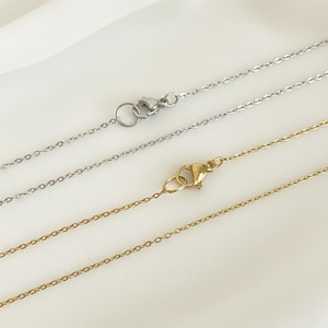 May include: Close-up of three delicate necklaces. One is gold-toned, and two are silver-toned. Each necklace features a fine chain and a clasp. The chains are displayed against a white, slightly textured background.