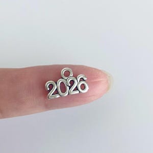 May include: A small, silver-colored charm with the number "2026" in a rounded font. The charm has a small loop at the top for attaching to a necklace or bracelet. The charm is shown against a light background.
