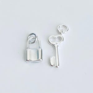 May include: A silver padlock and key charm set. The padlock has a square shape with a rounded top and bottom. The key has a traditional design with a heart-shaped bow.
