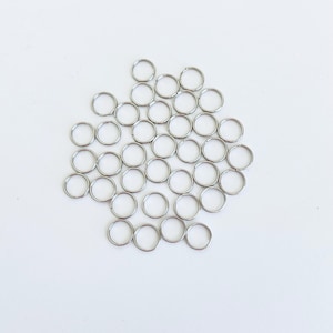May include: A large quantity of silver jump rings, arranged in a circular pattern on a white background.
