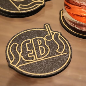 Puede incluir: Un posavasos redondo negro con un contorno dorado y la palabra "SEB'S" en una fuente estilizada.