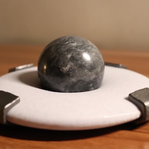 Puede incluir: Objeto decorativo con una esfera de mármol gris sobre una base circular blanca. La base tiene cuatro acentos cuadrados de color gris oscuro. El objeto se exhibe sobre una superficie de madera.