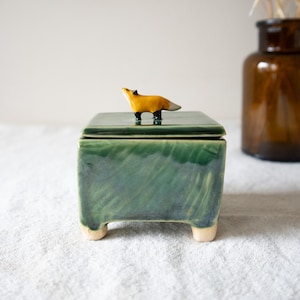Puede incluir: Caja de cerámica artesanal con acabado verde brillante y una pequeña figura de zorro amarillo. La caja tiene forma cuadrada y se apoya en cuatro pequeños pies de color claro. Artículo decorativo.