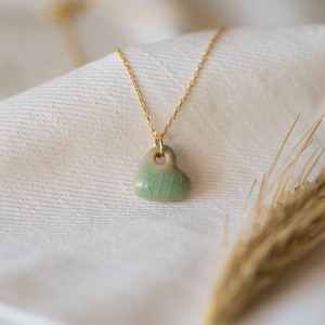 May include: A delicate gold chain necklace with a small, green ceramic heart pendant. The heart has a subtle, textured surface.