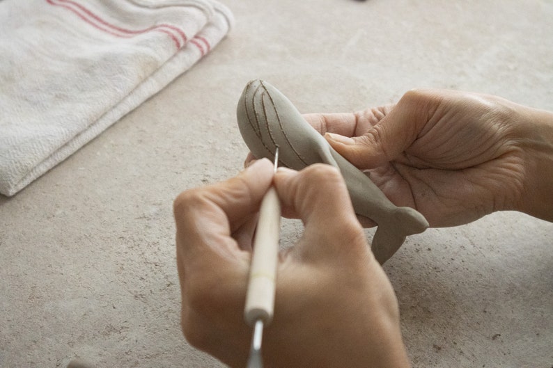 May include: A person's hands are sculpting a gray clay whale with a tool. The whale has a detailed design on its back.