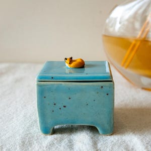 May include: A small, blue ceramic box with a lid. The box has a sleeping fox figurine on top of the lid. The box has four small feet.