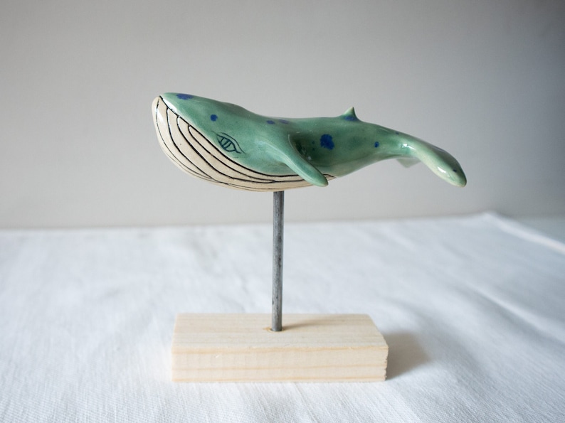May include: A ceramic whale sculpture with blue and green glaze, white stripes, and blue dots. The whale is mounted on a wooden base with a metal rod.