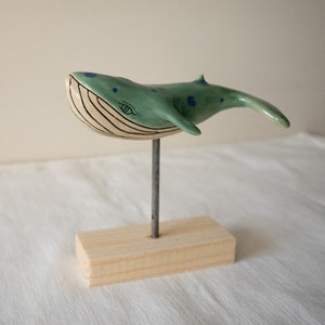 May include: A ceramic whale sculpture with blue and green paint, a white belly, and a black eye. The whale is mounted on a wooden stand.