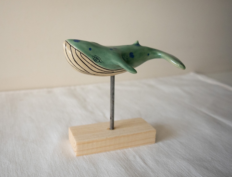 May include: A ceramic whale sculpture with blue and green paint, a white belly, and a black eye. The whale is mounted on a wooden stand.