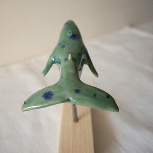 May include: A ceramic whale sculpture with blue and green speckled paint. The whale is in a swimming position with its tail raised.