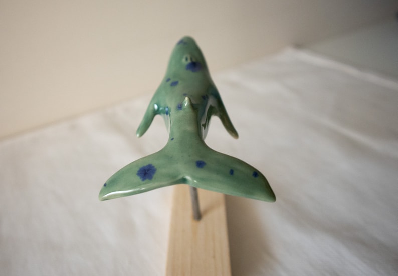 May include: A ceramic whale sculpture with blue and green speckled paint. The whale is in a swimming position with its tail raised.