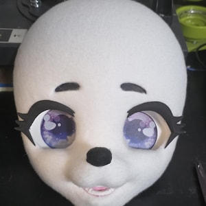 May include: A white, plush animal head with large, expressive eyes. The eyes have a purple and blue pattern, framed by black eyelashes and eyebrows. A black nose and a pink mouth complete the design, suggesting a friendly character.