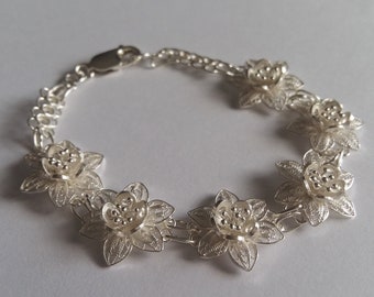 May include: A silver bracelet with a delicate filigree design featuring six flower-shaped charms. The bracelet is made of fine silver and has a polished finish.