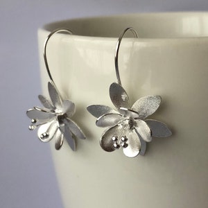 May include: Silver flower earrings with a delicate design. The earrings are made of sterling silver and have a brushed finish.