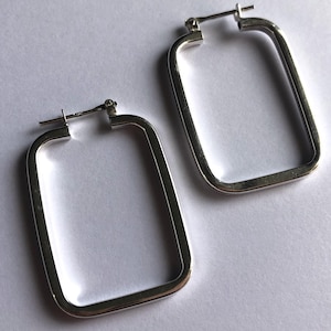 Könnte beinhalten: Zwei silberne, rechteckige Creolen. Die Ohrringe haben ein schlichtes, modernes Design mit rechteckiger Form. Die Ohrringe werden auf einer weißen Oberfläche gezeigt. Die Ohrringe sind aus glänzendem Metall gefertigt.