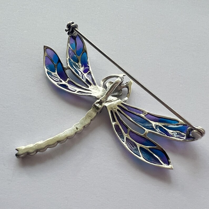 Pode incluir: Broche de lib&eacute;lula prateada com asas esmaltadas em azul e roxo. O broche tem um fecho de alfinete na parte de tr&aacute;s. O corpo da lib&eacute;lula &eacute; detalhado com um design texturizado. Um acess&oacute;rio decorativo.