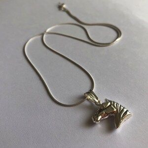 Puede incluir: Un collar de cadena de plata con un colgante de cabeza de caballo de plata.