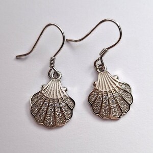May include: A pair of silver seashell earrings with a hook closure. Each earring features a detailed shell design, embellished with small, sparkling stones. The earrings are displayed against a white background.