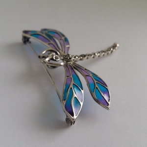 Pode incluir: Broche de lib&eacute;lula prateada com asas esmaltadas em azul e roxo. O corpo da lib&eacute;lula &eacute; detalhado com pequenos detalhes brilhantes. As asas s&atilde;o divididas em sec&ccedil;&otilde;es de azul claro e roxo.