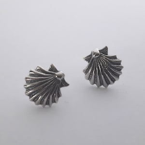 Può includere: Un paio di orecchini a lobo a forma di conchiglia argentata. Ogni orecchino presenta un design dettagliato a forma di ventaglio con creste strutturate e una piccola sommità arrotondata. Gli orecchini sono su uno sfondo bianco.