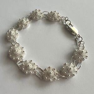 Puede incluir: Un brazalete de plata con una serie de cuentas de filigrana esféricas y ornamentadas, conectadas por delicados eslabones. El brazalete tiene un cierre seguro y se muestra sobre un fondo blanco. El intrincado diseño muestra una artesanía detallada.