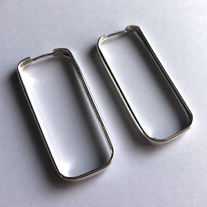May include: A pair of silver-toned hoop earrings featuring a rectangular shape with rounded corners. The earrings have a smooth, polished metallic finish and a thin wire post mechanism at the top for securing the hoops in place.