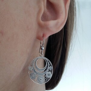 Puede incluir: Pendientes de filigrana de plata con un dise&ntilde;o circular. Los pendientes cuelgan de la oreja de una persona.