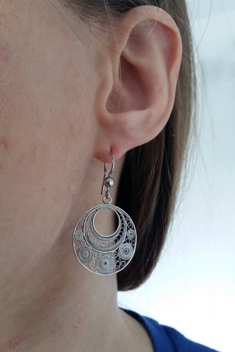 Puede incluir: Pendientes de filigrana de plata con un dise&ntilde;o circular. Los pendientes cuelgan de la oreja de una persona.