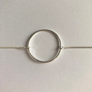 Puede incluir: Un collar plateado con una fina cadena y un colgante circular. El colgante es un anillo pulido, suspendido de la cadena. El collar se presenta sobre un fondo blanco, destacando su diseño minimalista y su elegancia sencilla.