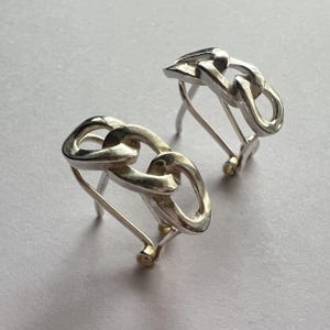 May include: A pair of silver-toned chain link earrings. Each earring features three interlocking links and a post-and-clasp closure. The earrings are displayed on a light gray surface.