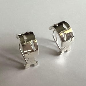 May include: A pair of silver-toned clip-on earrings. Each earring has a curved, rectangular shape with cut-out geometric designs. The earrings are displayed on a white surface, showcasing their reflective, polished finish.