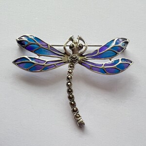 Pode incluir: Broche de lib&eacute;lula prateada com asas esmaltadas em azul e roxo. O corpo da lib&eacute;lula &eacute; detalhado com pequenas pedras redondas prateadas. As asas apresentam um efeito gradiente.