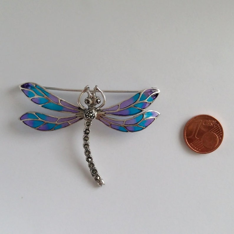 Pode incluir: Broche de lib&eacute;lula prateada com asas esmaltadas em azul e roxo. O corpo da lib&eacute;lula &eacute; detalhado com pedras de marcasite. Uma moeda de euro est&aacute; ao lado do broche para compara&ccedil;&atilde;o de tamanho.