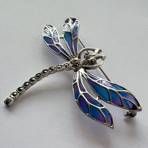 Pode incluir: Broche de lib&eacute;lula prateada com asas esmaltadas em azul e roxo. O corpo da lib&eacute;lula &eacute; detalhado com pedras de marcasite. As asas apresentam um design em camadas com tons de azul e roxo. O broche tem um fecho de alfinete.