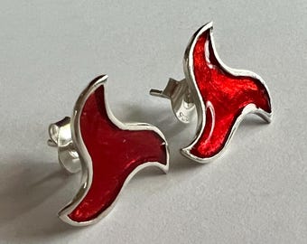May include: A pair of sterling silver stud earrings. Each earring features a red enamel design with a curved, flame-like shape. The earrings have a polished finish and are set against a light background.