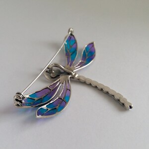 Pode incluir: Broche de lib&eacute;lula prateada com asas esmaltadas azuis e roxas. As asas t&ecirc;m um efeito de vitral e o corpo &eacute; detalhado. O broche tem um fecho de alfinete na parte de tr&aacute;s.