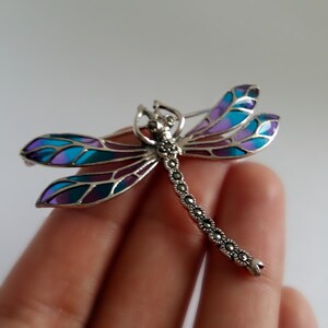 Pode incluir: Broche de lib&eacute;lula prateada com detalhes em marcasite e asas esmaltadas coloridas. As asas apresentam tons de azul, roxo e verde-azulado. Um acess&oacute;rio decorativo.