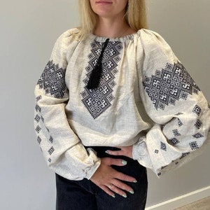 May include: A white linen blouse with black embroidery featuring a geometric pattern. The blouse has a round neckline and long sleeves.