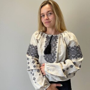 May include: A woman wearing a beige linen blouse with black embroidered geometric patterns. The blouse has a tassel detail at the neckline.