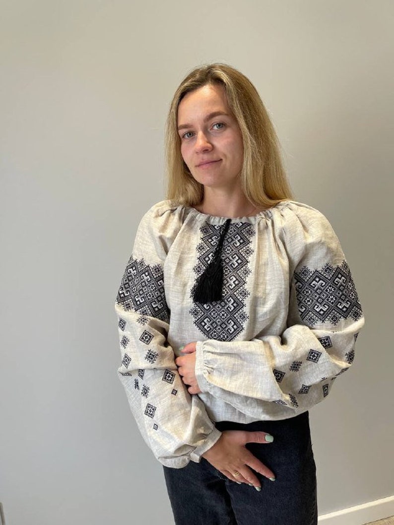 May include: A woman wearing a beige linen blouse with black embroidered geometric patterns. The blouse has a tassel detail at the neckline.