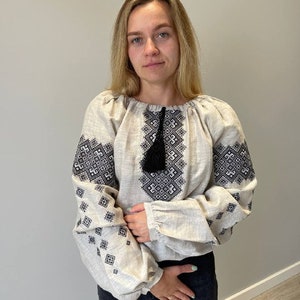 May include: A white linen blouse with black embroidered geometric patterns. The blouse has a gathered neckline and long sleeves with a black tassel.